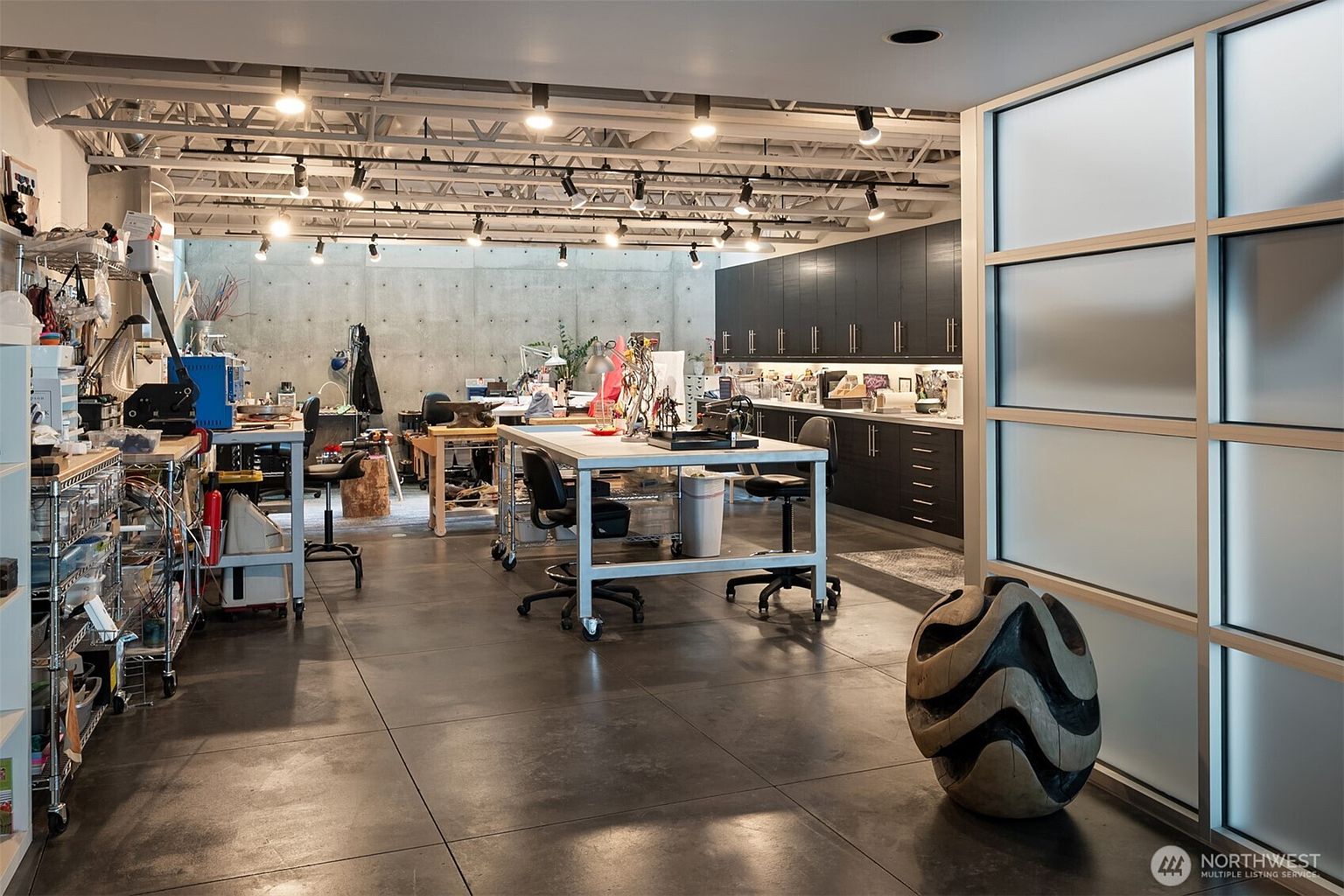 This spacious, industrial-style studio features a raw concrete wall, exposed ceiling joists, and dark cabinetry, creating a professional and creative workspace. The room is equipped with multiple work tables, rolling chairs, and extensive track lighting, making it ideal for artistic or technical projects. A large, frosted glass partition wall separates the space, while a unique sculptural piece sits in the foreground, adding a modern artistic touch to the concrete-floored environment.
