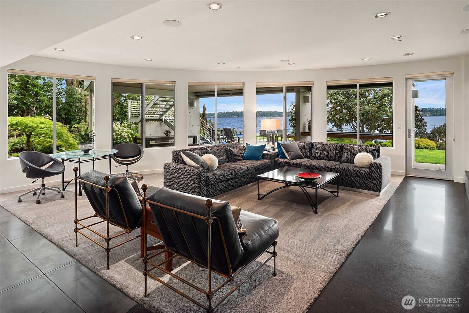 This spacious living room features a large, curved sectional sofa and two leather armchairs arranged on a neutral area rug. The room is defined by a series of large windows that provide expansive views of the lake and outdoor landscape, creating a bright and airy atmosphere. A glass-topped dining table with modern chairs sits to the left, while the dark flooring provides a sleek contrast to the light-colored walls.