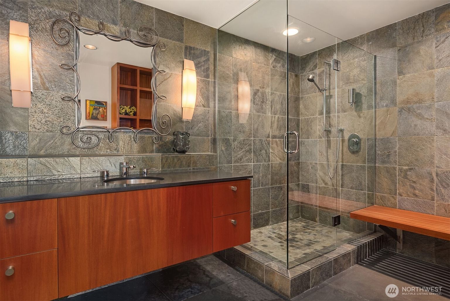 This modern bathroom features a floating wood vanity with a dark countertop and a decorative, ornate-framed mirror flanked by vertical wall sconces. A spacious walk-in shower is enclosed by glass panels and finished with textured stone wall tiles and a mosaic tile floor. A wooden bench extends from inside the shower to the exterior, creating a cohesive and spa-like aesthetic.