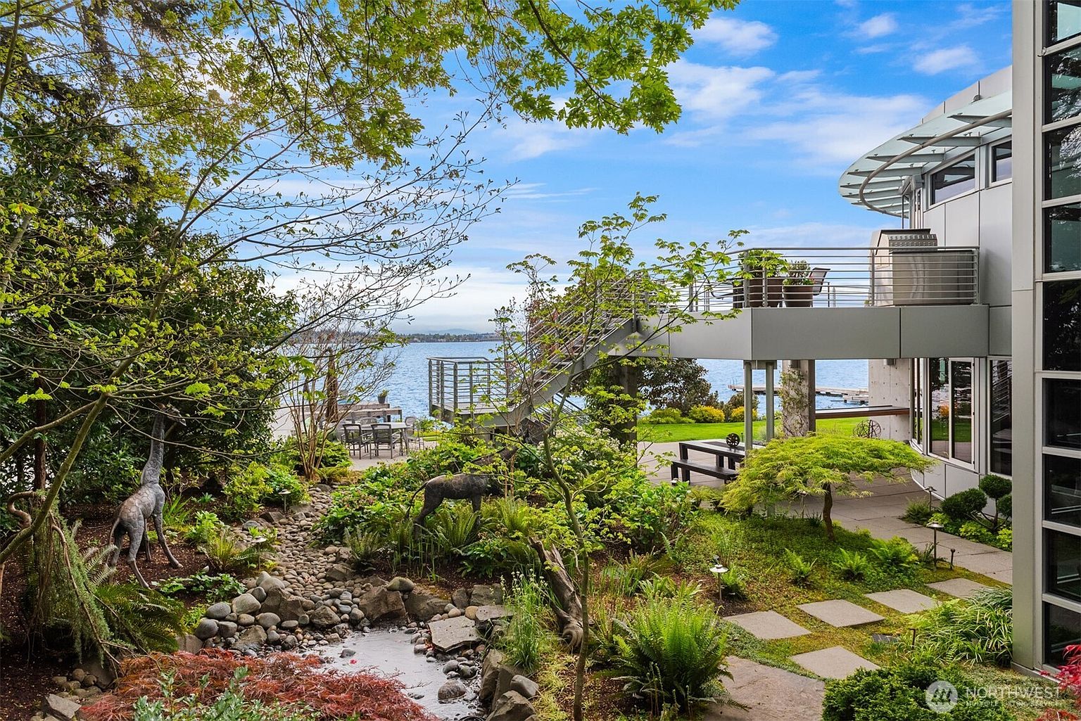 This contemporary waterfront property features a lush, landscaped garden with a stone-lined water feature and whimsical animal sculptures. A modern metal staircase connects the ground-level patio to an elevated deck, offering expansive views of the lake. The architecture incorporates large glass panels and clean lines, seamlessly blending the sophisticated structure with the natural surroundings.