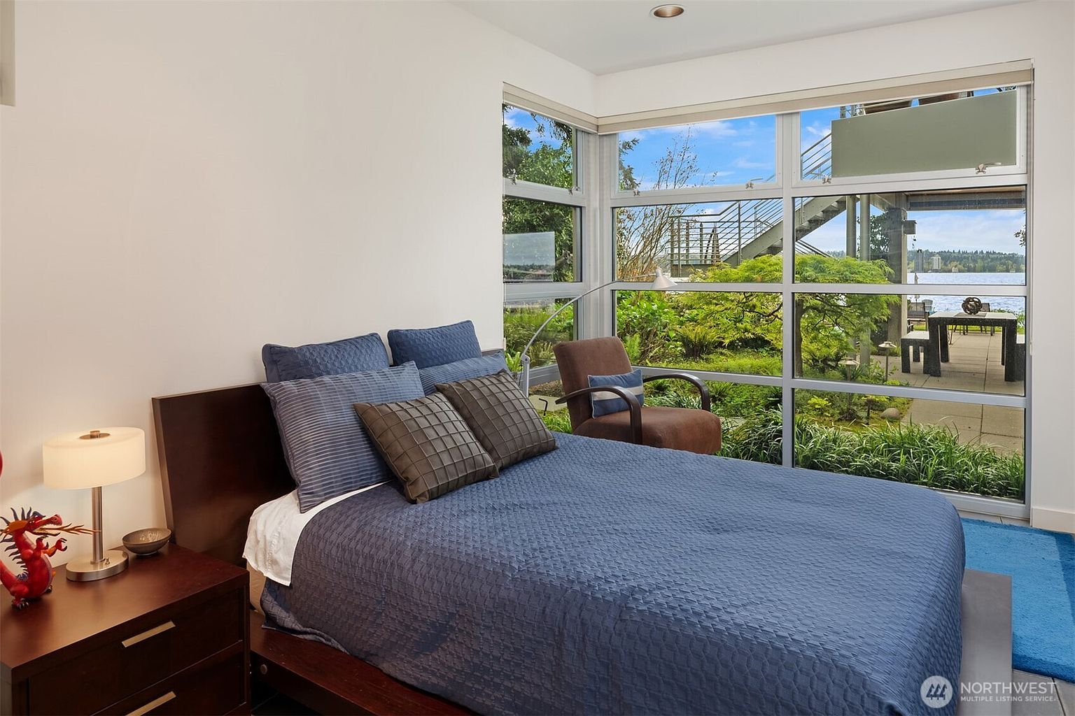 This bedroom features a contemporary design with a dark wood bed frame and a blue quilted comforter. Large windows provide an expansive view of the lush outdoor greenery and the waterfront beyond, creating a bright and serene atmosphere. A small brown armchair sits near the window, offering a cozy spot to enjoy the natural light and scenery.