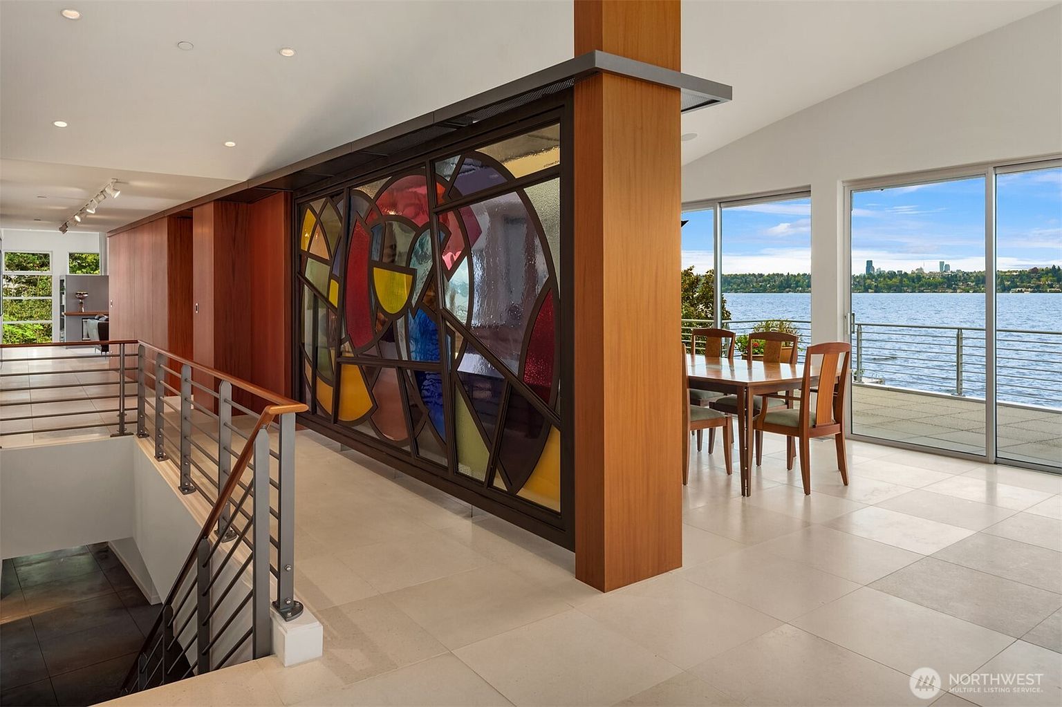This interior view captures a dining area adjacent to a staircase, featuring a striking stained-glass partition that serves as a focal point. The space is defined by light-colored tile flooring, a warm wood-paneled column, and large windows that provide an expansive view of the water and distant city skyline. The perspective looks from the landing toward the dining table, emphasizing the open, airy layout and the blend of artistic architectural elements with natural scenery.