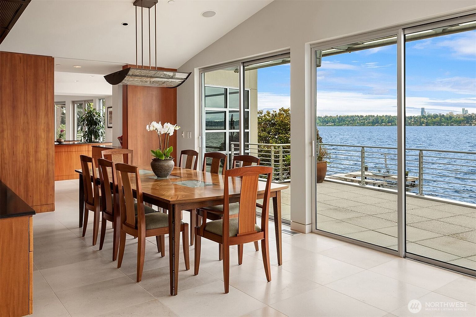 This bright dining room features a large wooden table surrounded by six chairs, positioned under a modern, rectangular pendant light. The space is defined by light-colored tile flooring and expansive sliding glass doors that provide a seamless transition to an outdoor deck overlooking a lake. The room's open-concept design flows into a kitchen area, creating an airy and sophisticated atmosphere with a view of the distant city skyline.