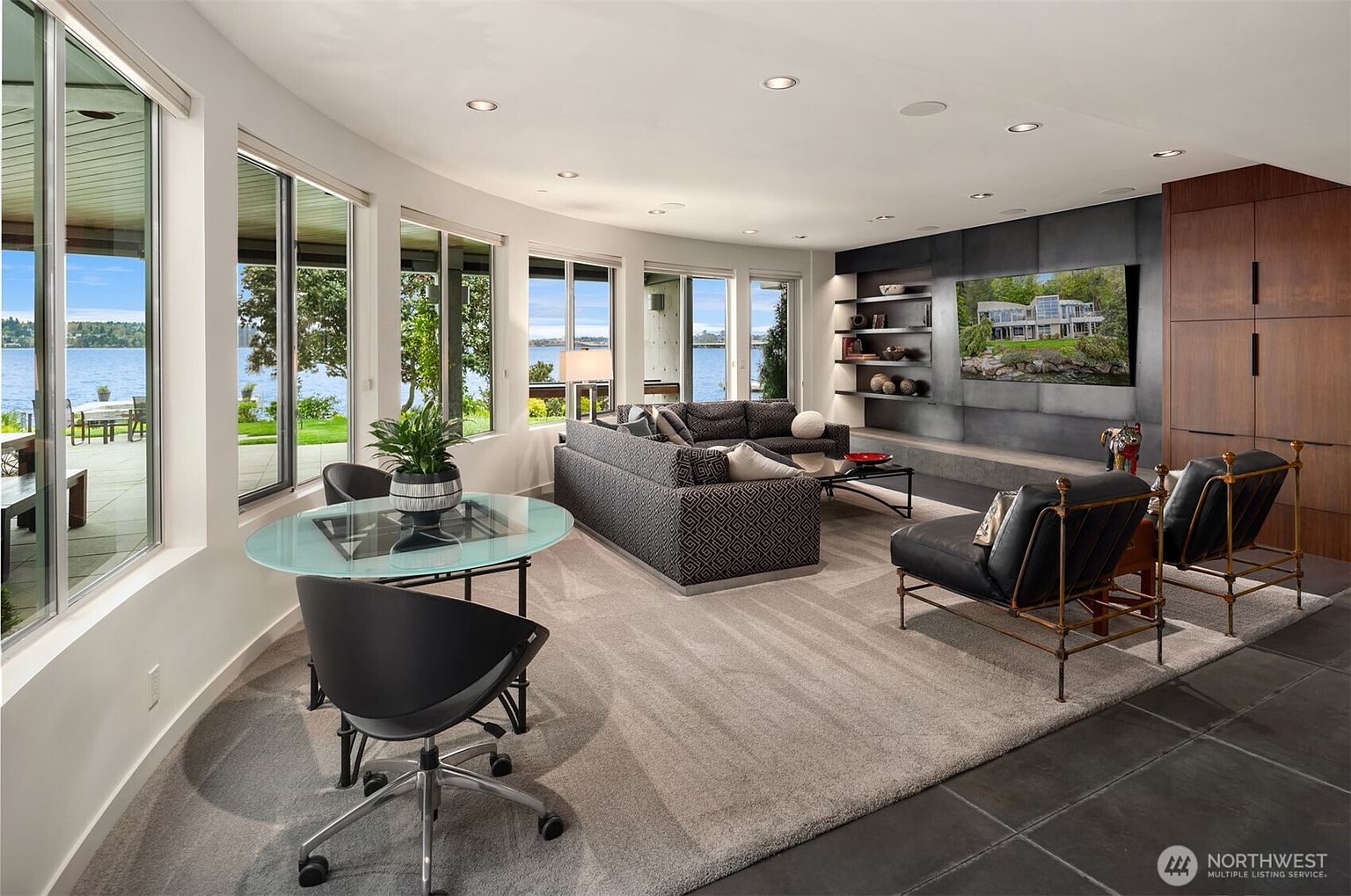 This spacious living room features a contemporary design with a large, patterned sectional sofa and two leather armchairs facing a wall-mounted television. A wall of large windows provides expansive views of the water and outdoor patio, filling the room with natural light. The space is anchored by a neutral area rug over dark flooring, with a small glass-topped dining table and chairs positioned near the windows.