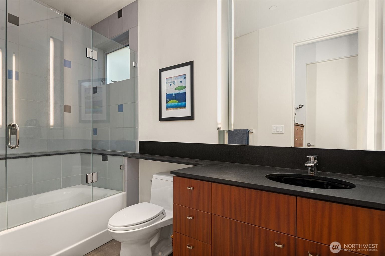 This modern bathroom features a sleek vanity with a dark stone countertop and warm-toned wood cabinetry. A glass-enclosed shower area with light-colored wall tiles sits adjacent to a white toilet, while a large mirror above the vanity enhances the sense of space. The room is accented by a framed piece of art on the wall, creating a clean and contemporary aesthetic.