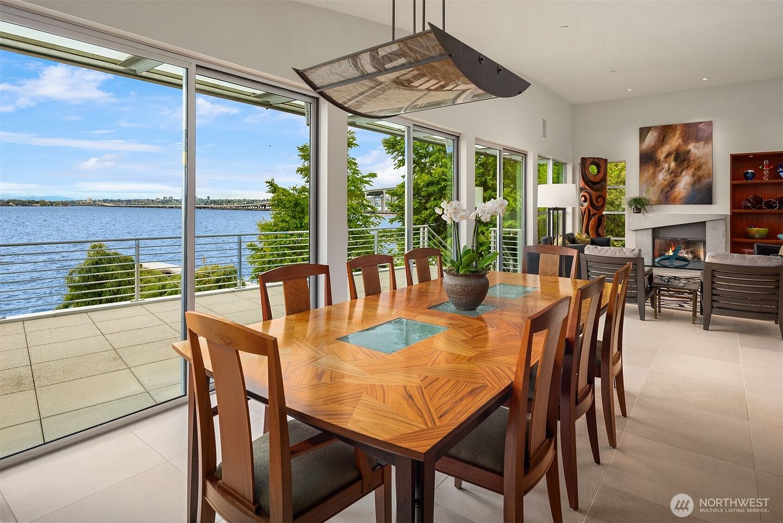 This bright dining area features a large wooden table with a geometric inlay pattern, surrounded by matching chairs. The room is defined by expansive glass sliding doors that provide a panoramic view of the water and bridge beyond, seamlessly connecting the interior to the outdoor patio. A modern, rectangular pendant light hangs above the table, while a cozy living area with a fireplace and a tall, carved wooden sculpture is visible in the background.