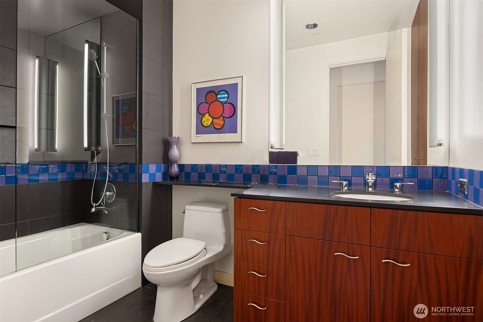 This contemporary bathroom features a dark wood vanity with a dark countertop and a white undermount sink. A glass-enclosed shower and tub combination is accented by a vibrant blue and purple tiled backsplash that extends along the wall. The space is decorated with a colorful floral art piece and includes a large mirror above the vanity, creating a bright and modern aesthetic.