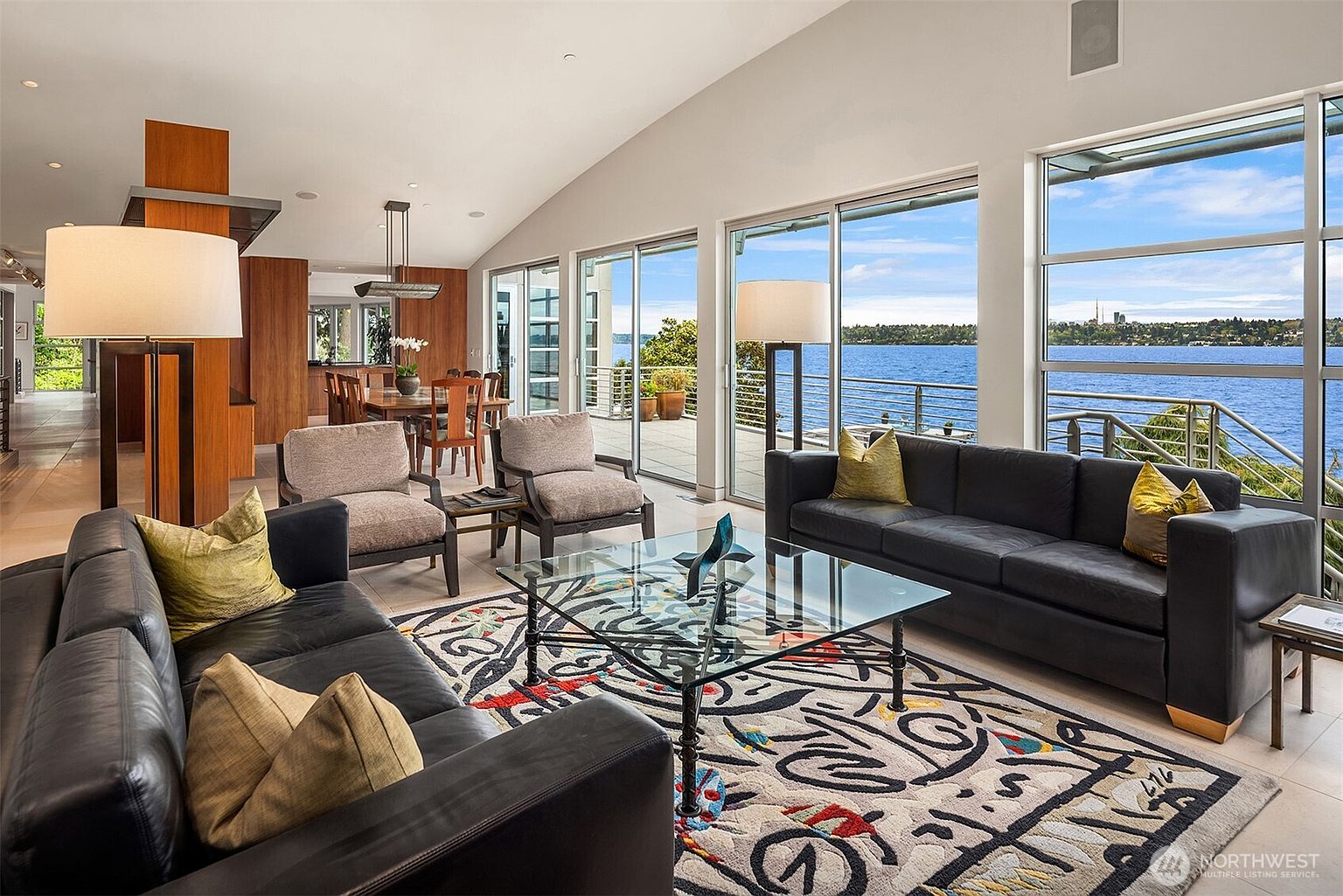 This spacious, open-concept living area features a contemporary design with dark leather sofas, a glass-topped coffee table, and a vibrant, patterned area rug. Large windows provide expansive views of the water and distant skyline, flooding the room with natural light. The space flows seamlessly into a dining area, creating an airy and sophisticated atmosphere perfect for entertaining.