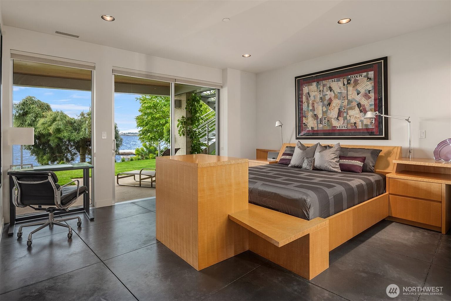 This modern primary bedroom features a sleek, light wood bed frame with an integrated footboard and matching side tables. Large sliding glass doors provide a seamless transition to an outdoor patio with water views, while dark tile flooring adds a sophisticated contrast to the neutral walls. A large, abstract framed artwork hangs above the bed, and a dedicated desk area is positioned near the window to take advantage of the natural light.