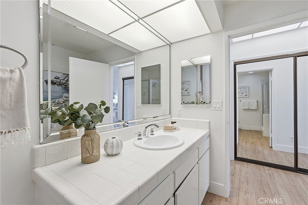This bright bathroom features a white tiled vanity countertop with an integrated sink and a large, expansive mirror that reflects the adjacent hallway and bedroom area. The space is accented by a decorative vase with greenery and a small pumpkin, while light-toned wood-look flooring extends throughout the room. A sliding mirrored door leads to an additional bathroom space, creating a sense of depth and openness in the layout.