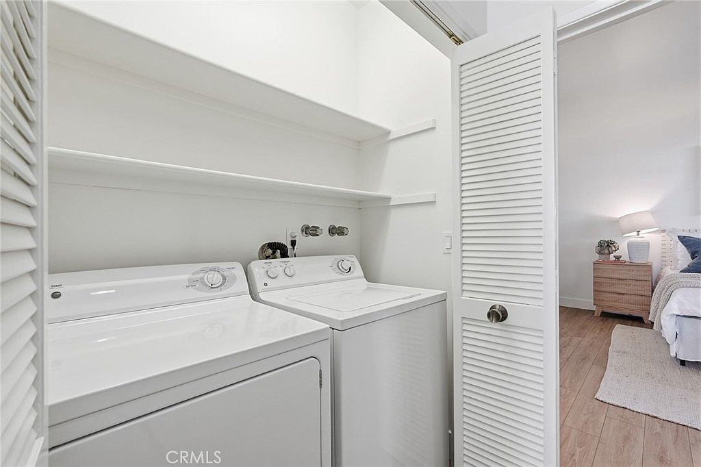 This laundry room features a side-by-side washer and dryer set positioned beneath two white wall-mounted shelves for storage. The space is enclosed by louvered bi-fold doors, with a view extending into an adjacent bedroom featuring light wood-look flooring and a bedside lamp. The room is bright and minimalist, offering a functional and compact utility area.