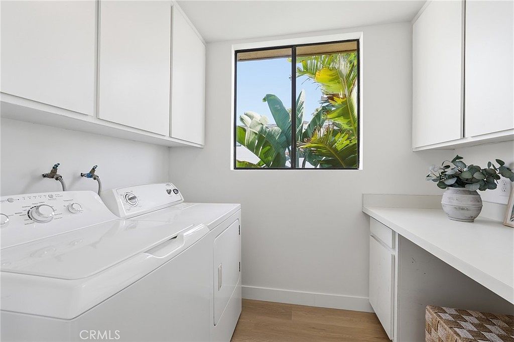This bright and functional laundry room features white cabinetry, a white washer and dryer set, and a long countertop providing ample workspace. A window above the appliances allows natural light to fill the room and offers a view of lush green foliage outside. The space is finished with light-toned flooring, creating a clean and organized atmosphere.