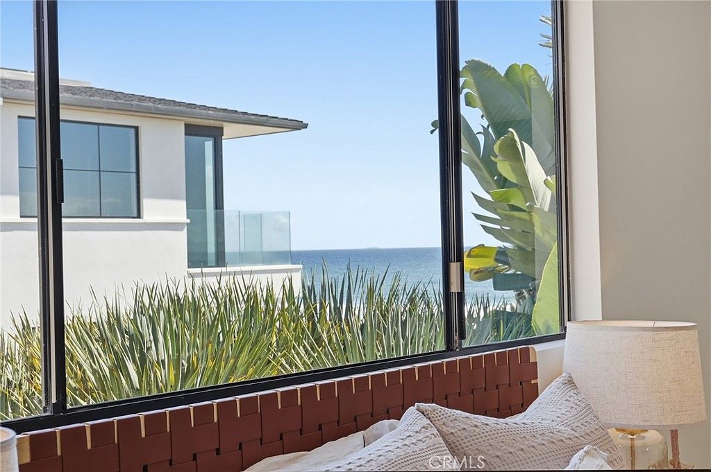 This interior view captures a serene bedroom setting, framed by large windows that offer a direct, picturesque view of the ocean and a neighboring modern home. In the foreground, a textured, woven-leather headboard and plush, patterned pillows create a cozy atmosphere, complemented by a bedside lamp with a neutral-toned shade. The perspective is intimate, emphasizing the seamless connection between the comfortable indoor living space and the expansive coastal scenery outside.