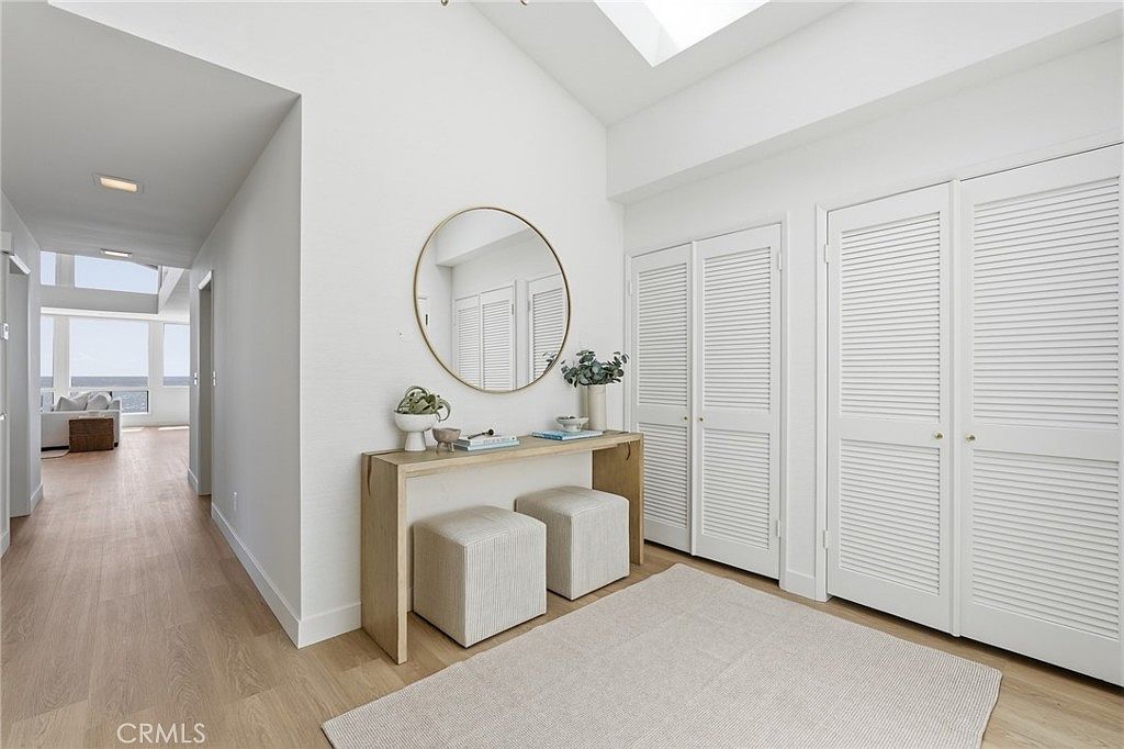 This bright and airy entryway features light wood flooring and crisp white walls, creating a clean, modern aesthetic. A console table with two matching ottomans sits beneath a large circular mirror, while louvered-door closets provide ample storage. The perspective looks down a hallway toward a sunlit living area with large windows, emphasizing the open and inviting flow of the home.