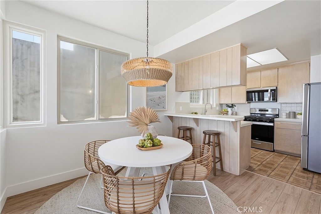 This bright dining area features a round white table surrounded by four woven chairs, set beneath a large, textured pendant light. The space flows directly into an open-concept kitchen with light wood cabinetry, a breakfast bar with two stools, and stainless steel appliances. Large windows provide ample natural light, creating an airy and inviting atmosphere with a cohesive, coastal-inspired aesthetic.