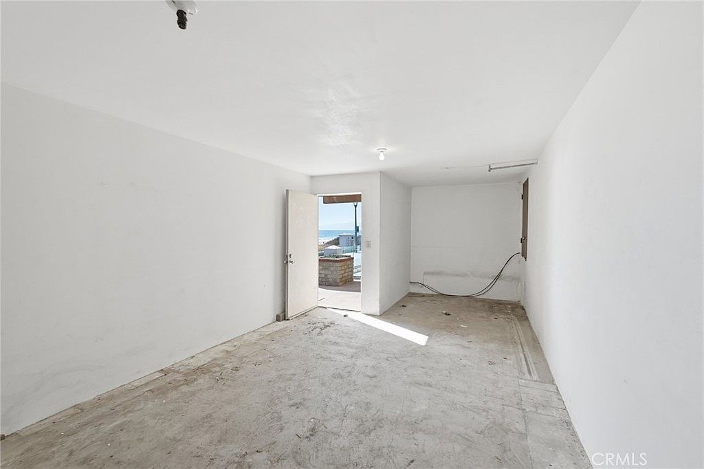 This image shows an unfinished, utilitarian room with plain white walls and a bare concrete floor. An open doorway at the far end provides a direct view of an outdoor patio and the ocean beyond, suggesting a beach-front property. The space appears to be a blank canvas, possibly a storage area, workshop, or a room undergoing renovation.