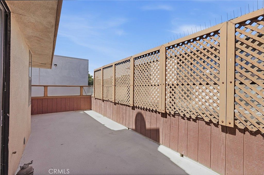 This image shows an outdoor balcony space featuring a concrete floor and a privacy screen made of wooden lattice panels. The balcony is enclosed by a combination of solid wood paneling and the lattice structure, which is topped with anti-bird spikes. The perspective is from the doorway looking out across the length of the balcony under a clear blue sky.