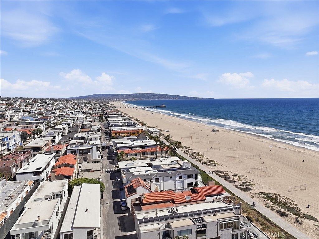 This high-angle aerial perspective captures a prime coastal neighborhood situated directly along a sandy beach. The scene features a dense collection of residential properties with varied rooflines and architectural styles, all oriented toward the expansive ocean view. A long pier extends into the water in the distance, emphasizing the desirable beachside location and the proximity to the shoreline.