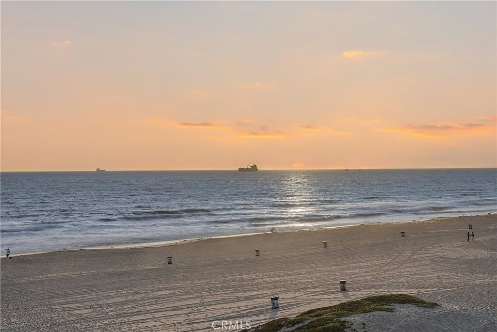 This elevated view captures a serene beach scene at sunset, likely taken from a balcony or terrace. The composition features a wide expanse of sandy beach meeting the ocean, with a large cargo ship visible on the horizon under a soft, pastel-colored sky. The perspective emphasizes the tranquil coastal location and the expansive horizon, highlighting the property's proximity to the water.
