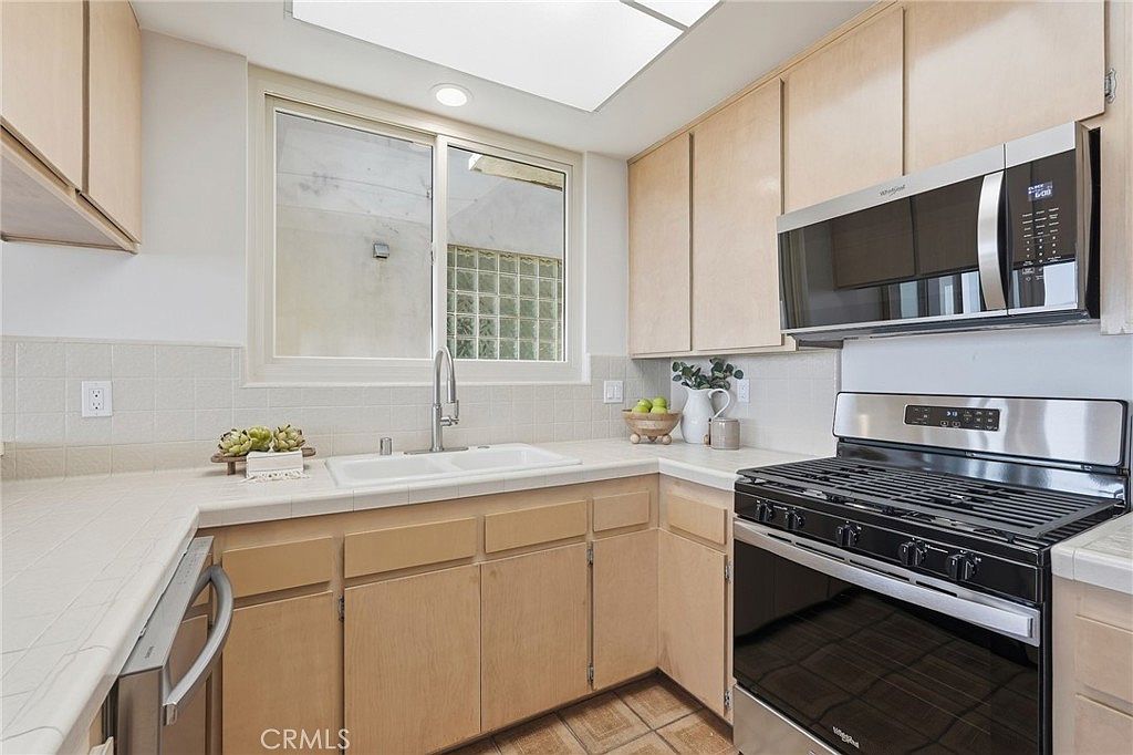 This kitchen features light wood cabinetry, a white tiled countertop, and stainless steel appliances, including a gas range and an over-the-range microwave. A large window above the double-basin sink allows natural light to brighten the space, which is accented by a decorative bowl of fruit and a small plant. The perspective is a straight-on view of the sink area, showcasing a functional and well-lit workspace.