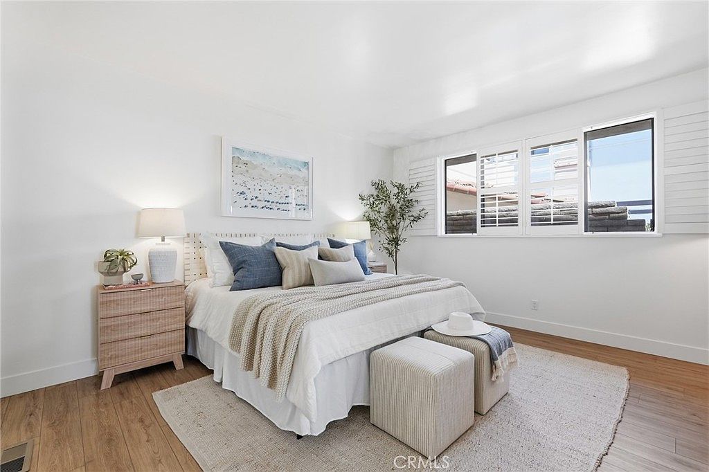 This bright and airy primary bedroom features a neutral color palette with light wood-tone flooring and crisp white walls. A large bed with layered bedding and decorative pillows serves as the focal point, flanked by matching nightstands and lamps. The room is completed by a textured area rug, two upholstered ottomans at the foot of the bed, and large windows with white shutters that allow for ample natural light.