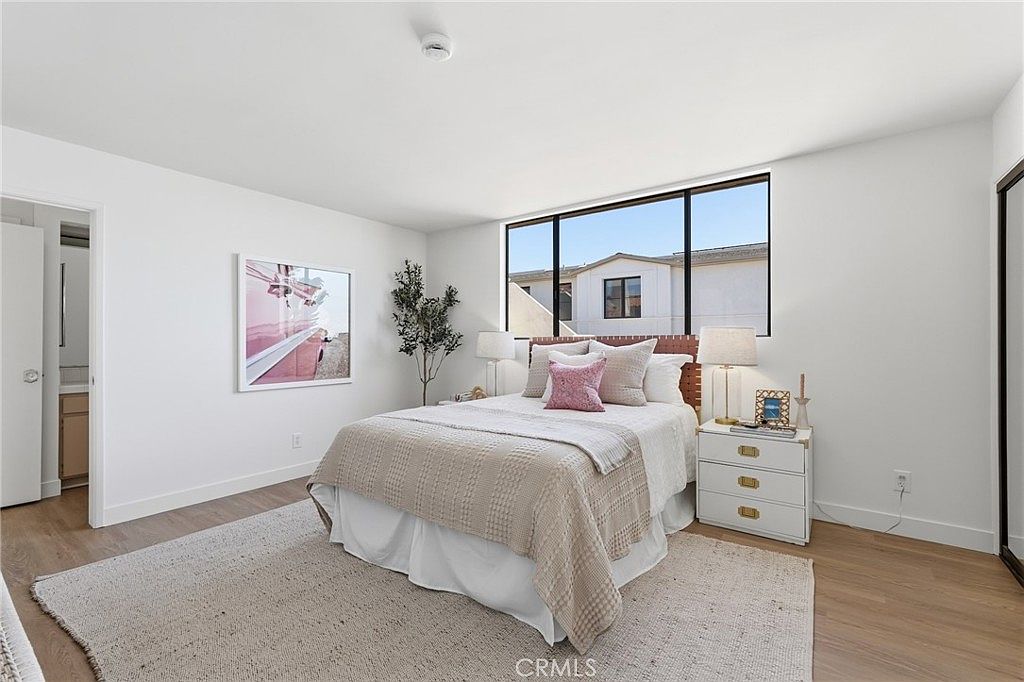 This bright and airy bedroom features light-toned wood flooring and crisp white walls, creating a clean and inviting atmosphere. A central bed with a textured headboard is dressed in neutral linens and accented with a pink throw pillow, while a matching white nightstand sits to the right. A large window provides natural light and a view of the neighboring building, and a framed piece of art hangs on the wall to the left.