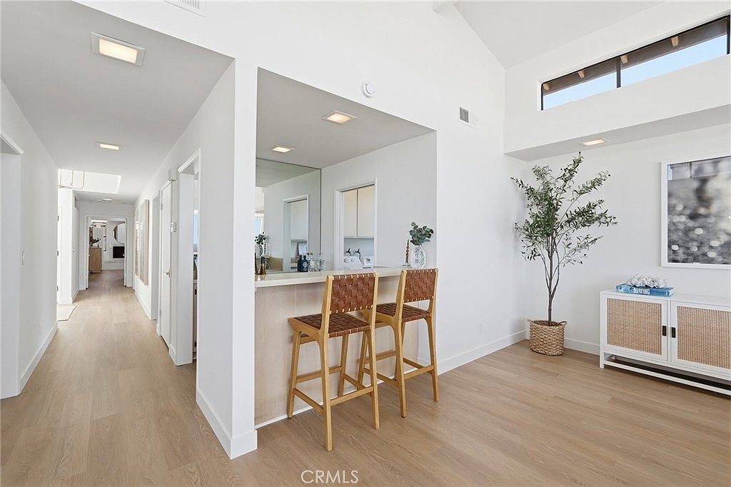 This bright, open-concept interior space features a hallway leading to other rooms, adjacent to a built-in bar area with two woven-back bar stools. The room is characterized by light wood-toned flooring, crisp white walls, and a vaulted ceiling with a clerestory window that enhances the natural light. A decorative potted tree and a console table with coastal-inspired decor add a welcoming, modern aesthetic to the living area.