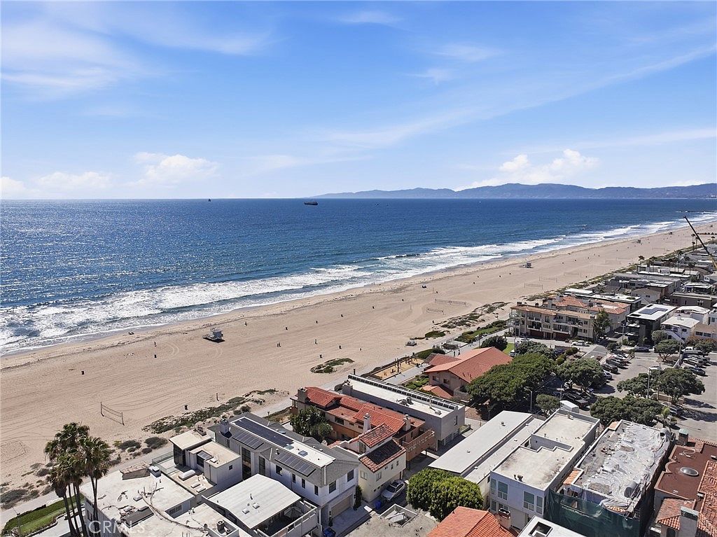 This high-angle aerial shot captures a prime coastal location, showcasing a stretch of sandy beach meeting the Pacific Ocean under a clear blue sky. In the foreground, a cluster of residential properties with varied architectural styles and rooftop decks sits directly adjacent to the shoreline. The perspective emphasizes the proximity of the homes to the water, highlighting the desirable beachside lifestyle.