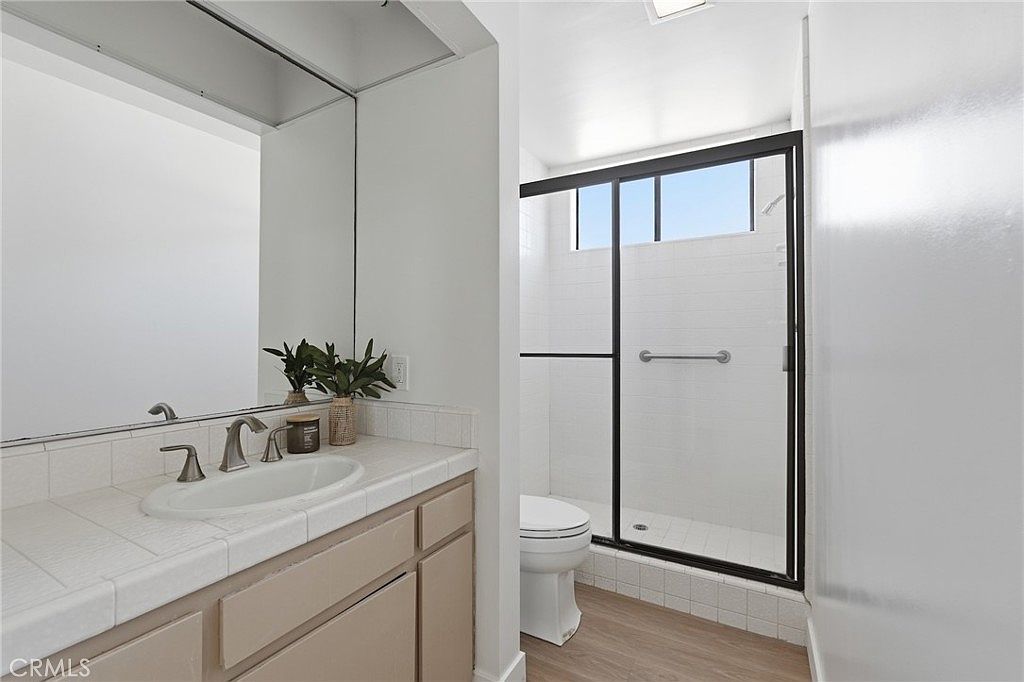 This bathroom features a vanity with a light-toned countertop and a built-in sink, complemented by a large wall-mounted mirror. To the right, a walk-in shower is enclosed by a black-framed glass door and features white wall tiles. The space is finished with light wood-look flooring, creating a clean and functional aesthetic.