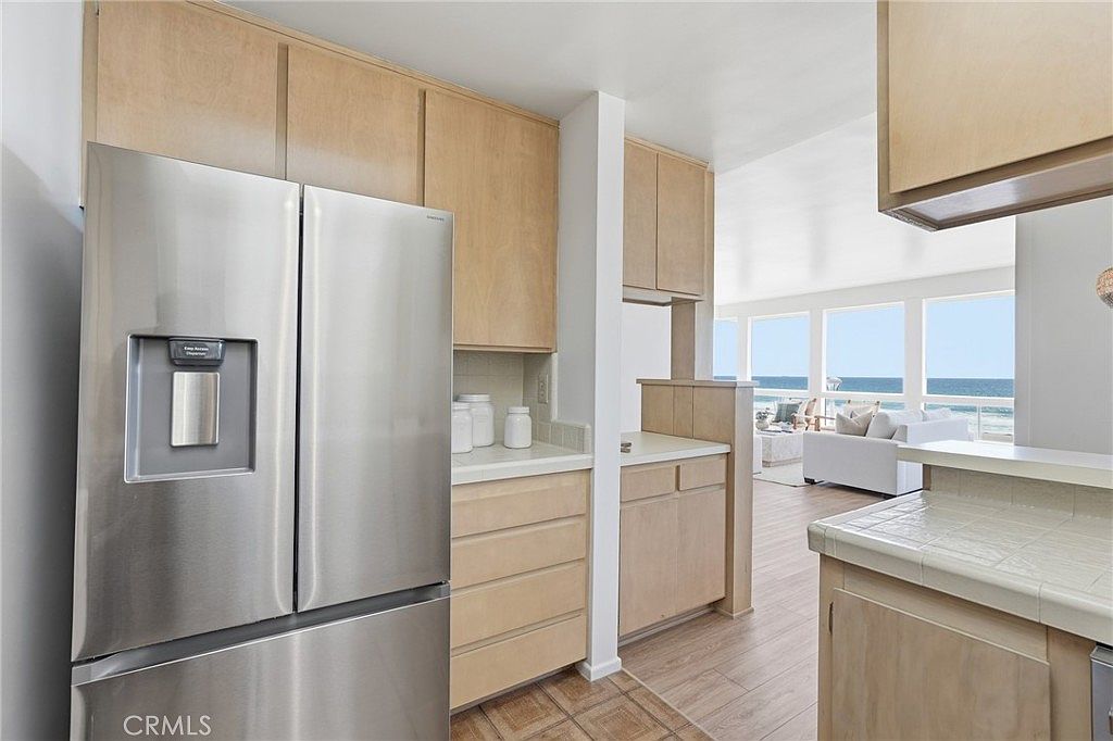 This kitchen features light wood cabinetry and a stainless steel refrigerator, creating a bright and functional workspace. The layout opens directly into a sunlit living area that offers expansive views of the ocean through large windows. The space is characterized by a clean, coastal aesthetic with light-toned flooring that flows seamlessly between the kitchen and the living room.
