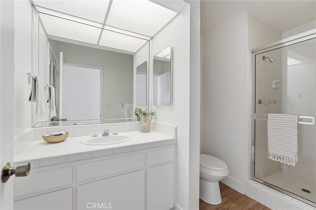 This bright, clean bathroom features a white vanity with a single sink and a large, full-width mirror that enhances the sense of space. To the right, a glass-enclosed shower with white tile walls provides a crisp, modern look, while the wood-toned flooring adds warmth to the neutral palette. The room is well-lit by a recessed ceiling light fixture, creating a welcoming and functional atmosphere.