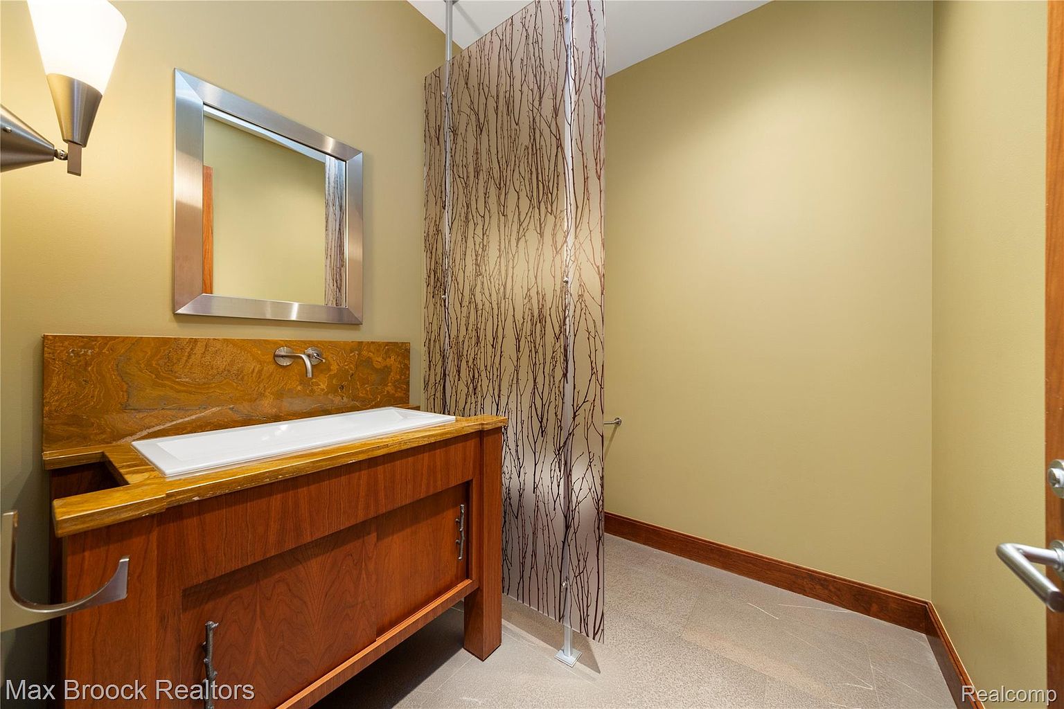 This is a bathroom featuring a wooden vanity with a white sink and a unique backsplash. A large mirror hangs above the sink, and a decorative glass partition separates the vanity area from the rest of the bathroom. The walls are painted in a neutral tone, and the flooring appears to be tile.