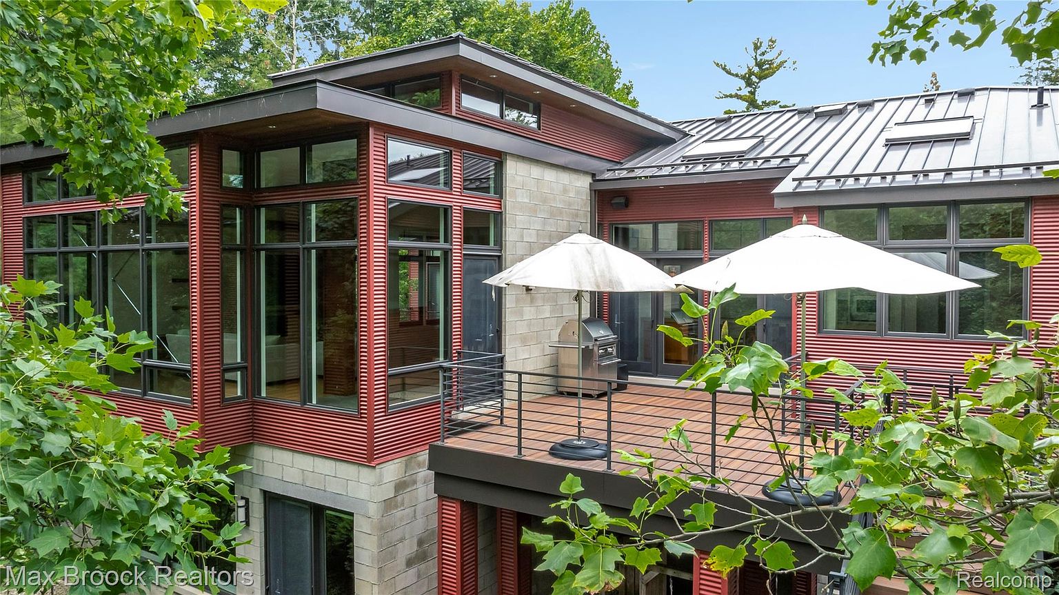 This image showcases a modern home's exterior, specifically highlighting a multi-level patio or deck area. The structure features a combination of materials, including red corrugated siding, concrete block, and dark-framed windows. The deck is furnished with a stainless steel grill and two white umbrellas, suggesting an outdoor living space perfect for entertaining.