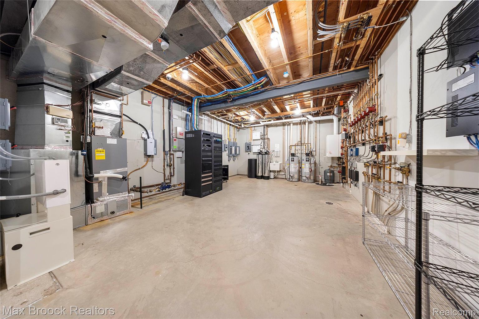 This is a spacious basement featuring exposed ductwork and ceiling joists, giving it an industrial feel. The room is equipped with various mechanical systems, including HVAC units, water heaters, and a server rack, suggesting a functional space. The concrete floor and neutral wall color create a clean, utilitarian aesthetic.