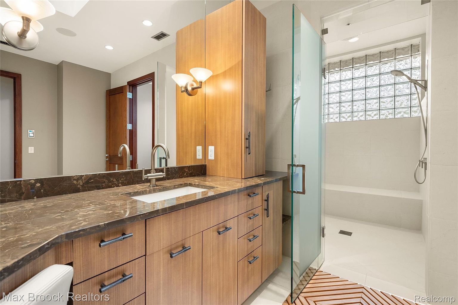 This is a well-lit primary bathroom featuring a modern vanity with a brown countertop and wooden cabinets. A large mirror reflects the vanity and a portion of the room, while a glass-enclosed shower with a built-in bench is visible to the right. The bathroom exudes a clean and contemporary aesthetic, with neutral tones and sleek fixtures.