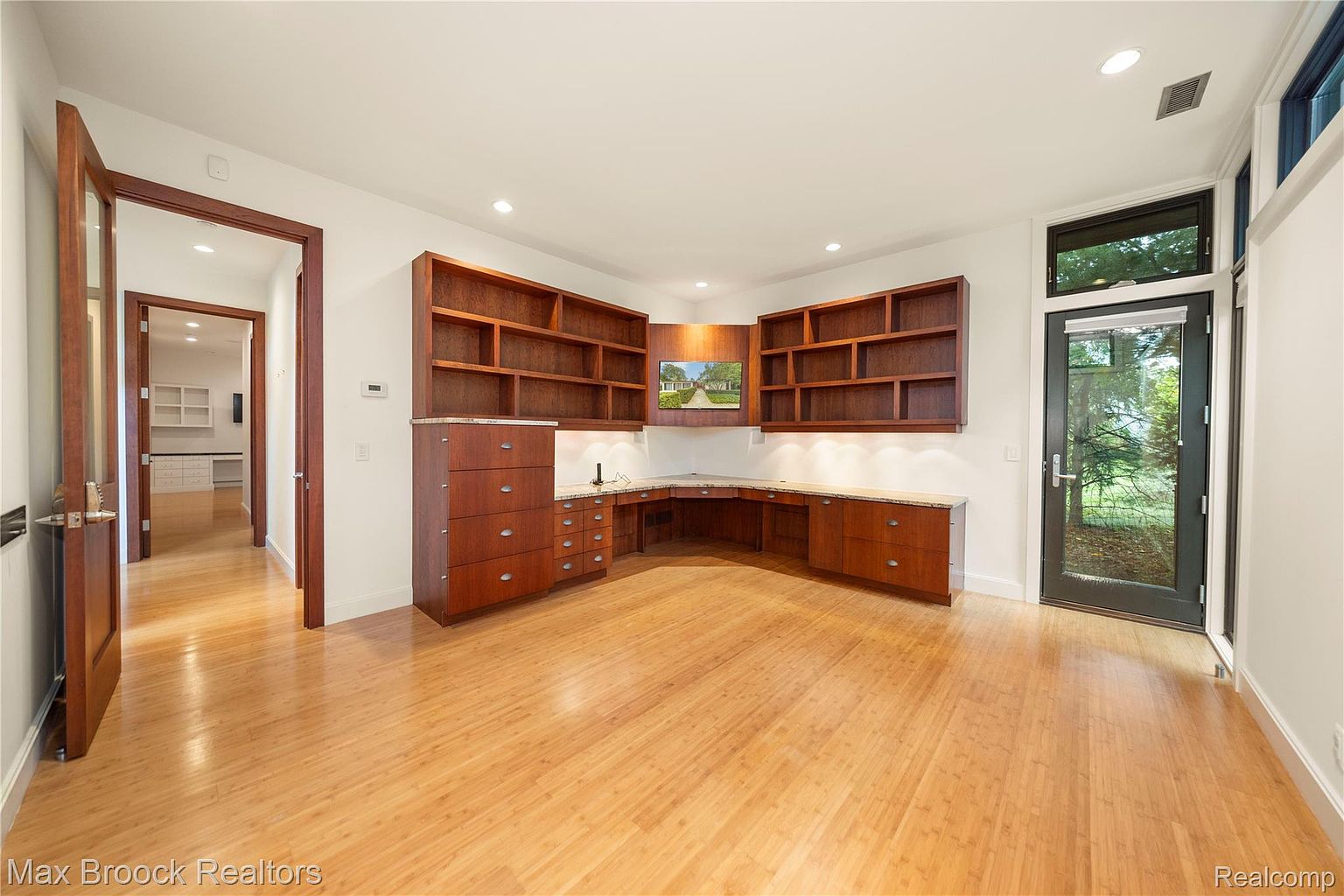 This is an interior shot of a home office featuring custom built-in wooden cabinetry and shelving. The room has hardwood floors and a door leading to the outside, providing natural light. The overall impression is a well-organized and functional workspace.