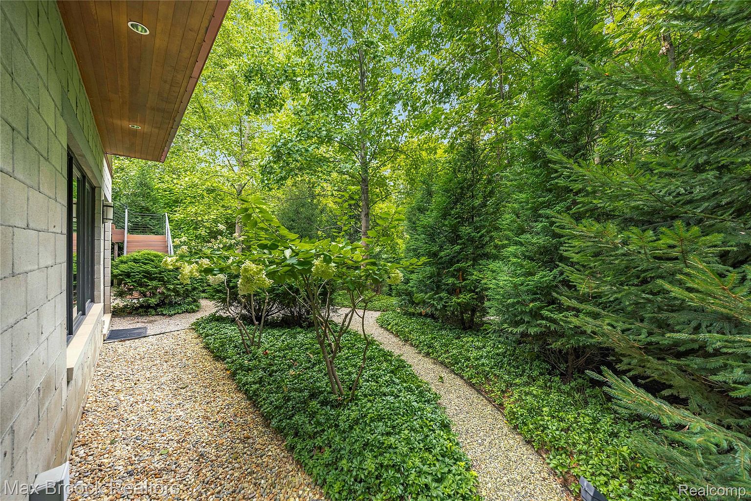 This image showcases a beautifully landscaped yard with winding gravel pathways bordered by lush greenery and mature trees. The garden features a variety of plants, including shrubs and ground cover, creating a serene and inviting outdoor space. The pathway leads away from the side of the house, suggesting a private and well-maintained property.