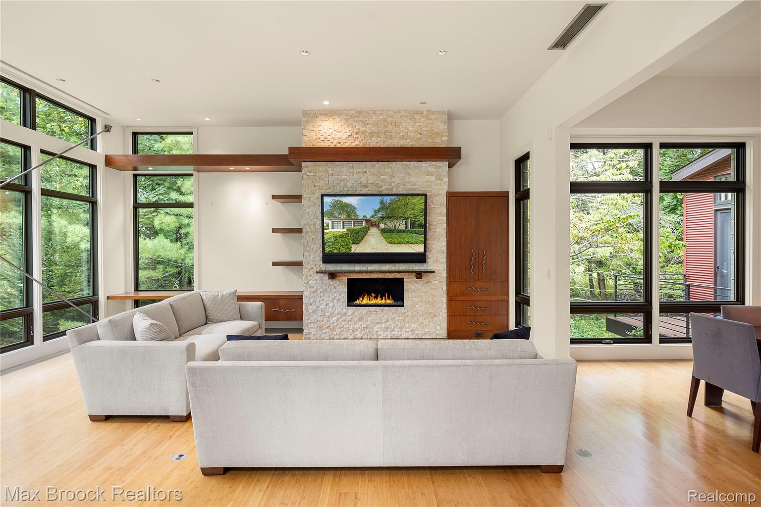 This is an interior shot of a modern living room featuring a stone fireplace with a mounted television and a built-in electric fireplace. The room is bright with natural light from large windows, and the hardwood floors add warmth. Two large sofas anchor the space, creating a comfortable and inviting atmosphere.