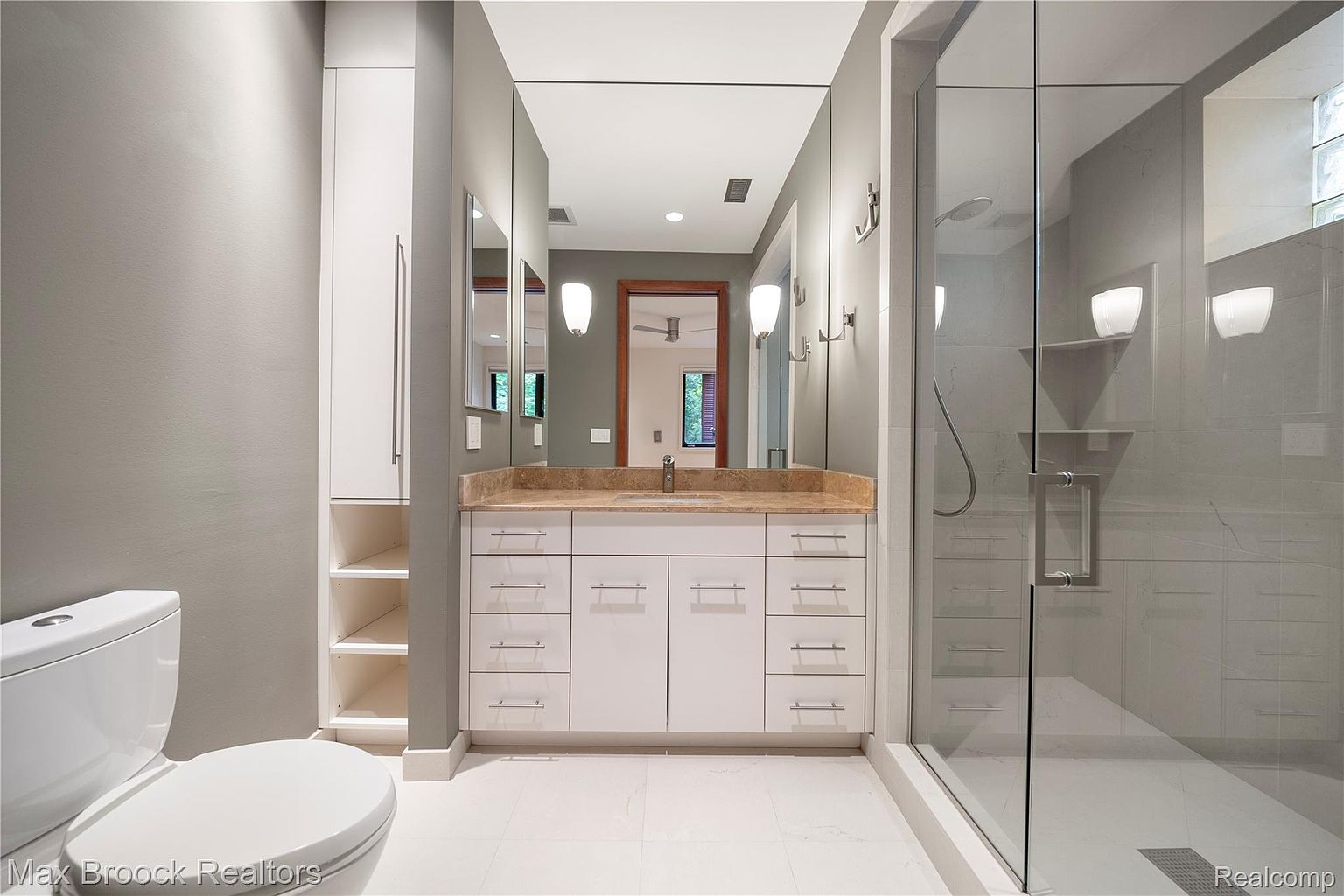 This is a well-lit bathroom featuring a white toilet, a vanity with white cabinets and a light brown countertop, and a large mirror. A glass-enclosed shower is visible on the right, showcasing light gray tiling and modern fixtures. The overall impression is clean, modern, and functional.
