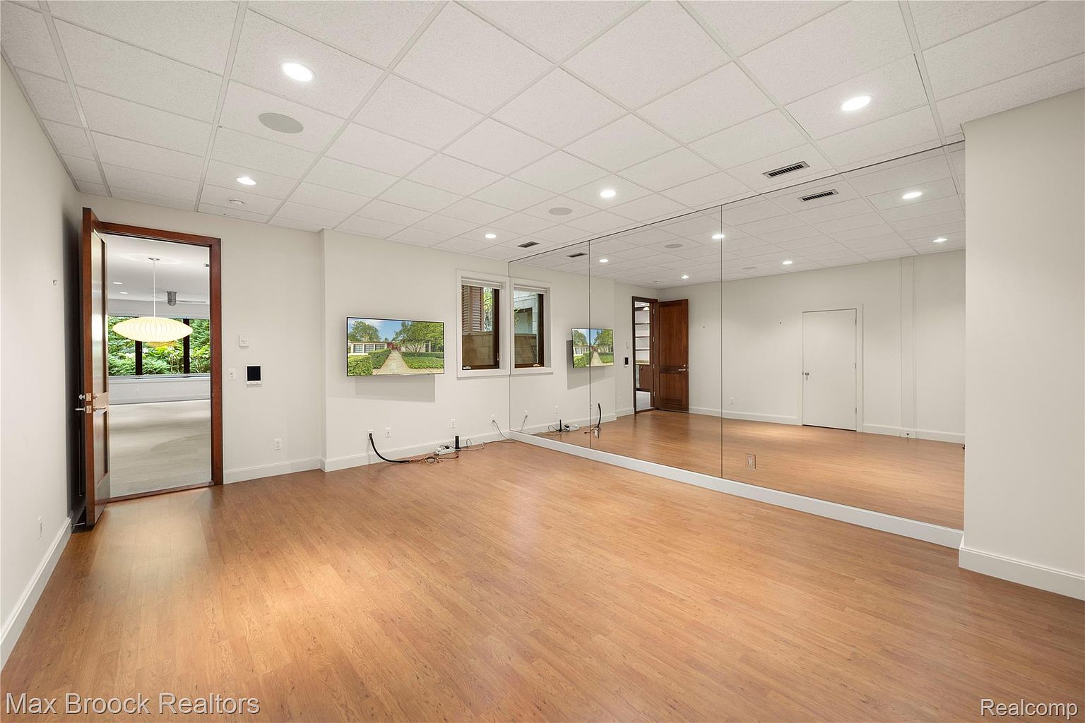 This is an interior shot of a home gym featuring wood flooring, a mirrored wall, and recessed lighting. Two flat-screen televisions are mounted on the wall, and there are two doors, one leading to another room and the other to a hallway. The room appears spacious and well-lit, suitable for various exercise activities.