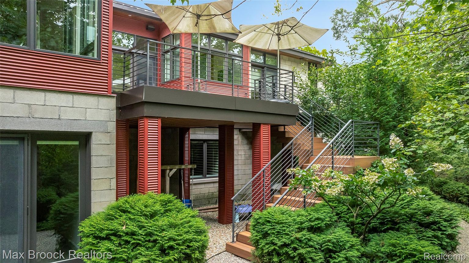 This image showcases the side of a modern home with a unique architectural design. The exterior features a combination of materials, including red corrugated metal, gray concrete blocks, and wood accents. A staircase leads to a second-story balcony with outdoor umbrellas, and lush greenery surrounds the property, creating a private and inviting atmosphere.