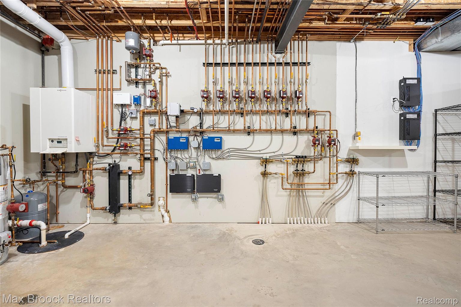 This is a basement utility room featuring extensive copper piping and mechanical systems mounted on a white wall. The concrete floor is bare, and metal shelving units stand to the side. The image showcases the complex infrastructure of the home, emphasizing its functionality.