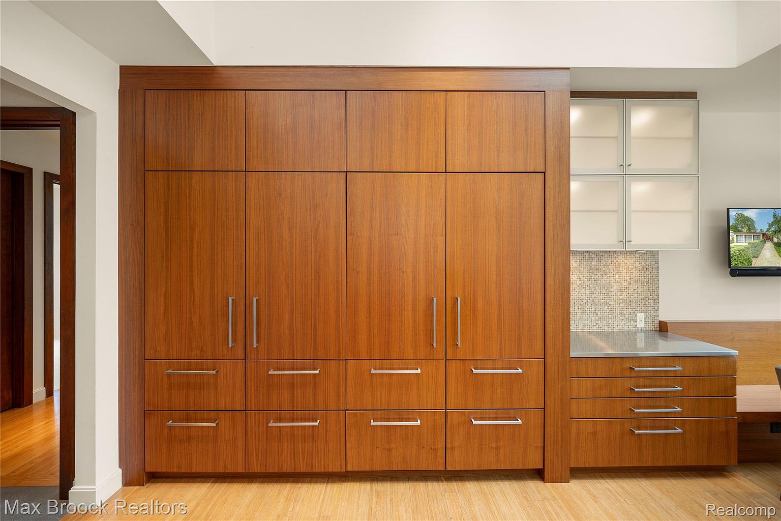 This is a well-organized kitchen featuring a large, built-in wooden cabinet system with sleek, modern hardware. The cabinets provide ample storage, and the adjacent countertop area includes additional drawers and cabinets, complemented by frosted glass upper cabinets. A small television is mounted on the wall, and the overall design is clean and contemporary.