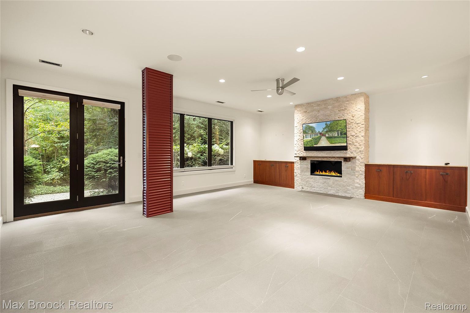 This is a spacious living room featuring a modern fireplace with a television mounted above it. The room has large windows offering views of the outdoors, and the flooring appears to be a light-colored tile. Built-in cabinetry lines the walls, providing ample storage space, and a ceiling fan adds to the room's comfort.