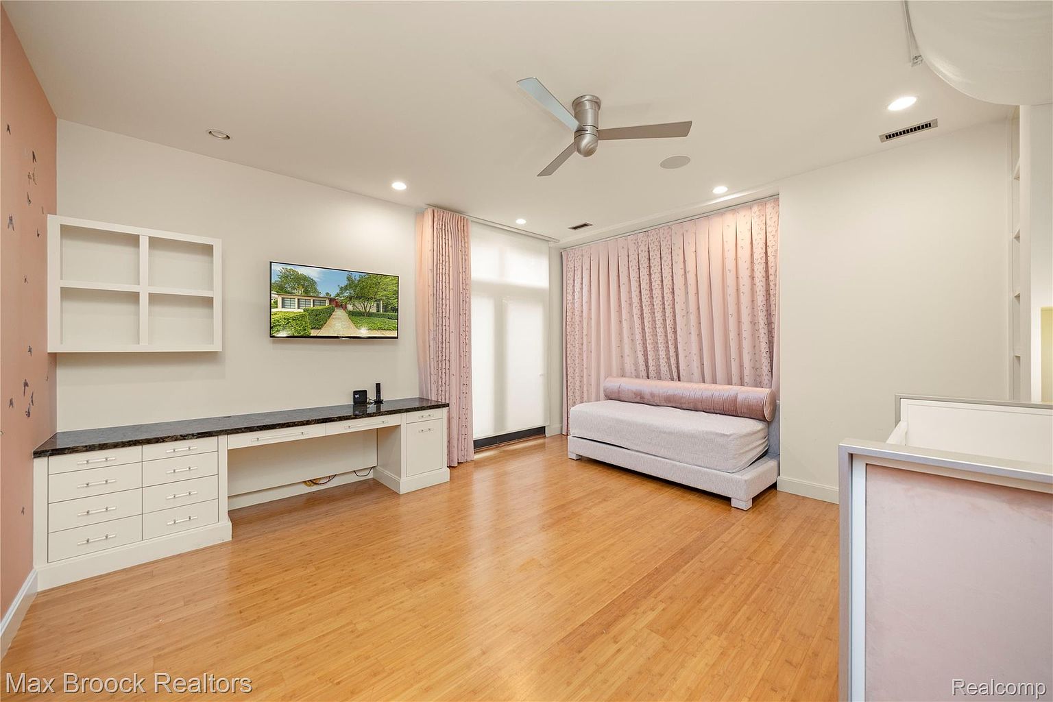 This is an interior shot of a bedroom featuring a built-in desk with drawers and a black countertop, a wall-mounted television, and a daybed with pink curtains. The room has hardwood floors and a ceiling fan, creating a bright and airy atmosphere. The overall style is modern and comfortable, suitable for a guest room or secondary bedroom.