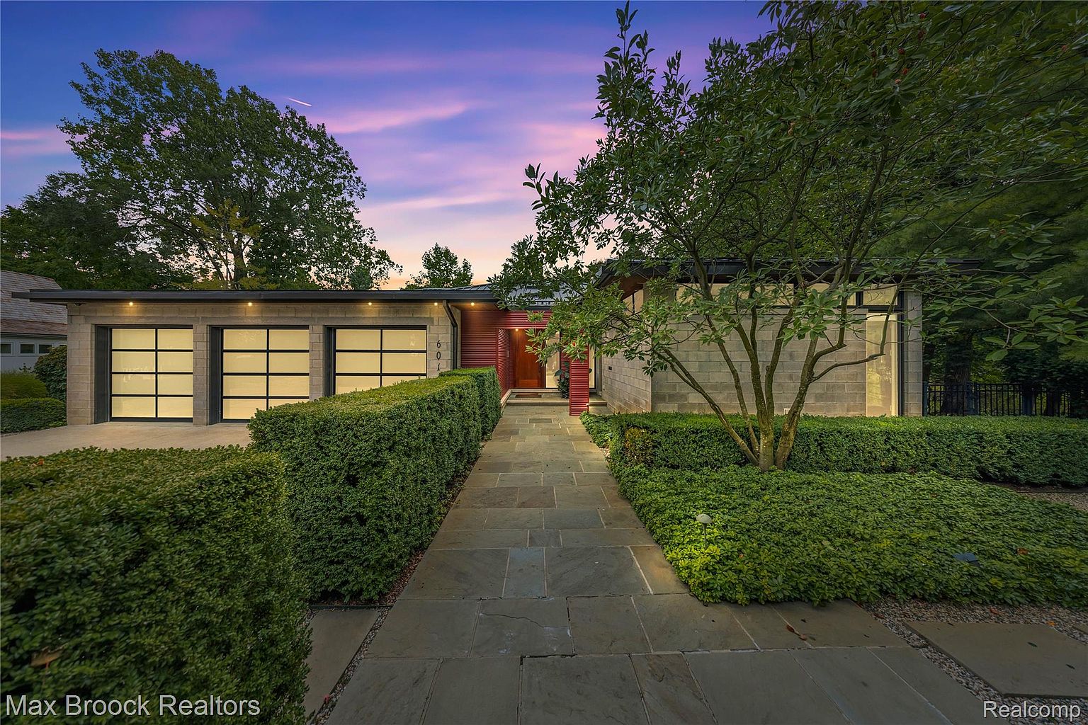 This is a front exterior view of a modern home with a well-manicured landscape. The house features a stone facade, a flat roof, and large garage doors with black frames. A stone pathway leads to a red-accented entryway, flanked by neatly trimmed hedges and a mature tree, creating a welcoming and sophisticated curb appeal.