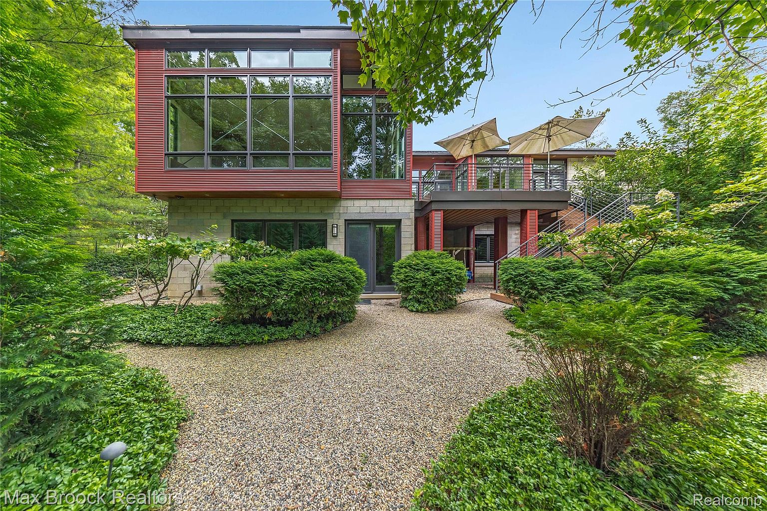 This is a front view of a modern, two-story home with a striking red and gray exterior. The house features large windows, a balcony with patio umbrellas, and a gravel driveway surrounded by lush greenery. The architectural style is contemporary, emphasizing clean lines and a connection to the surrounding landscape.