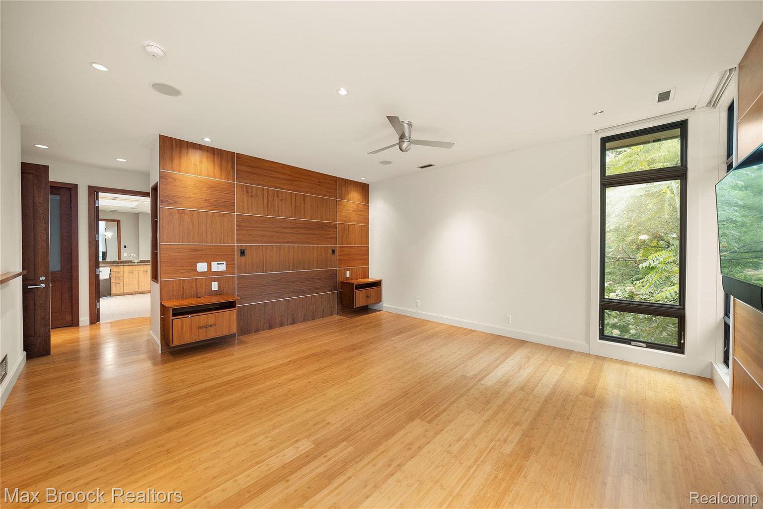 This is a spacious primary bedroom featuring hardwood floors and a unique wood-paneled accent wall with built-in nightstands. A large window provides natural light and a view of the outdoors. The room has a modern and minimalist aesthetic, creating a serene and inviting atmosphere.