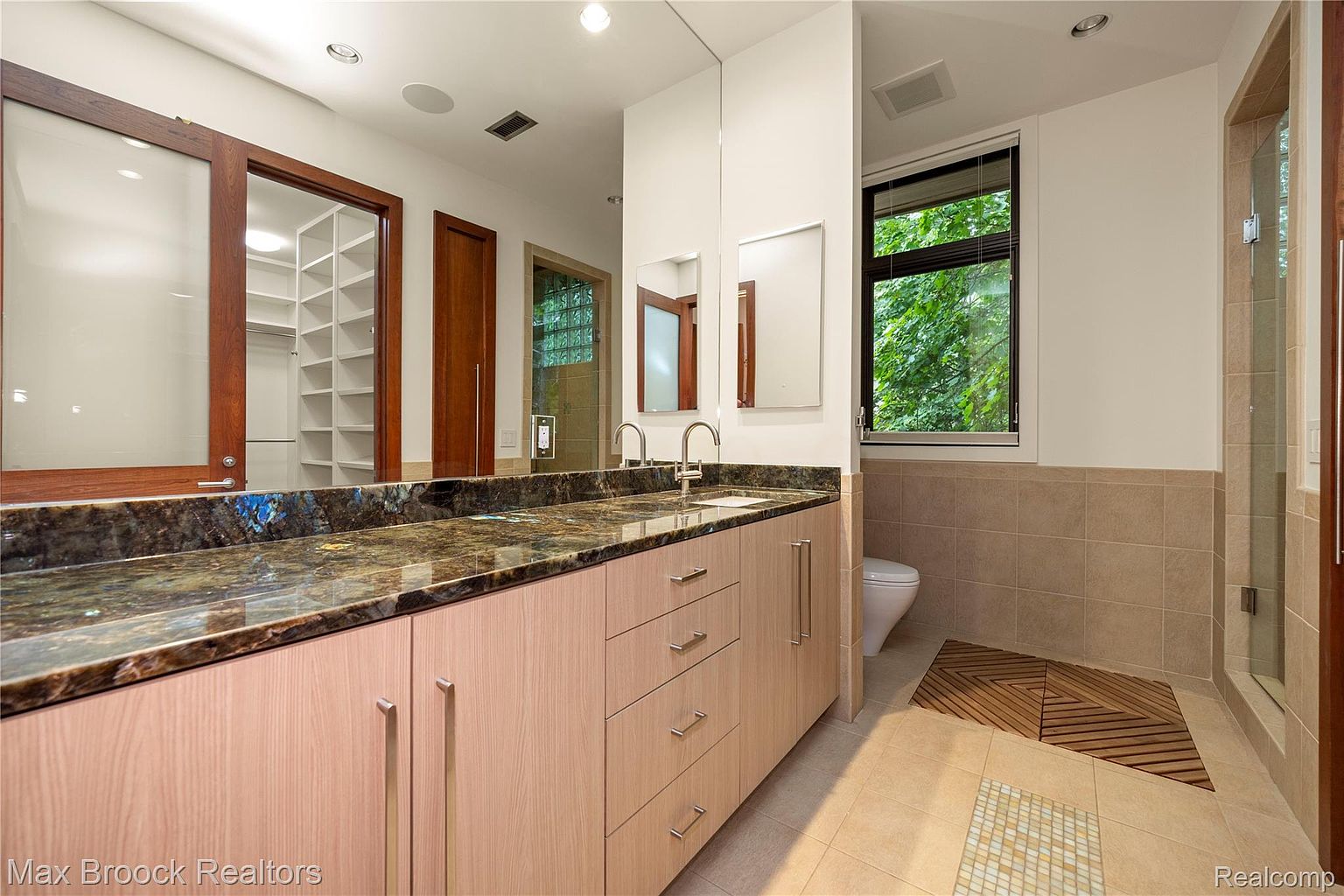 This is a primary bathroom featuring a long vanity with a dark granite countertop and light wood cabinets. A large mirror spans the length of the vanity, reflecting a walk-in closet and a glass-enclosed shower. The bathroom has neutral-toned tiles and a modern aesthetic.