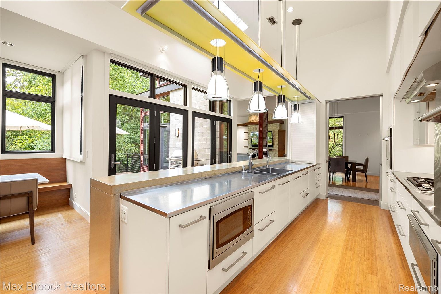 This is a bright, modern kitchen featuring sleek white cabinetry, stainless steel countertops, and appliances. The kitchen is well-lit with pendant lighting and natural light from the large windows and doors that offer a view to the outside. The open layout and clean lines create a contemporary and inviting space.